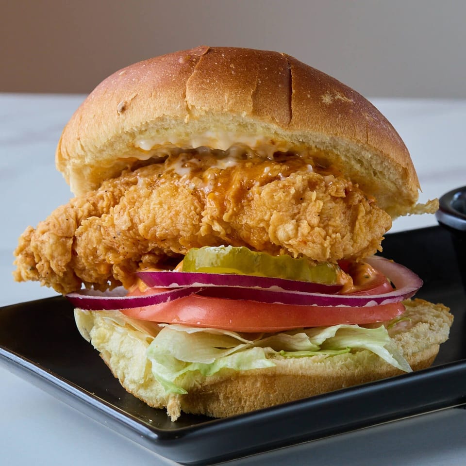 A Flaming Hot Halal Grill fried chicken sandwich with lettuce, tomato, onion, and pickles on a bun.